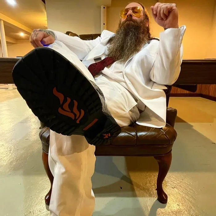 Reverend Meantooth in a white suit and amber glasses, boot raised toward the camera